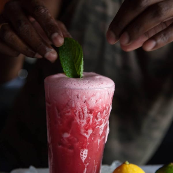 Hands garnishing a tall glass of foamy pink Caribbean drink with a fresh mint sprig.