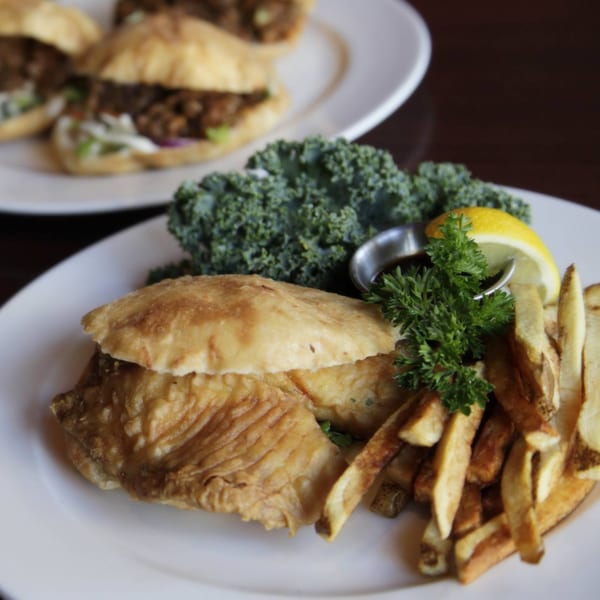 Fried fish sandwich with fries, kale, lemon, and parsley on a white plate at Pam's Caribbean Kitchen.